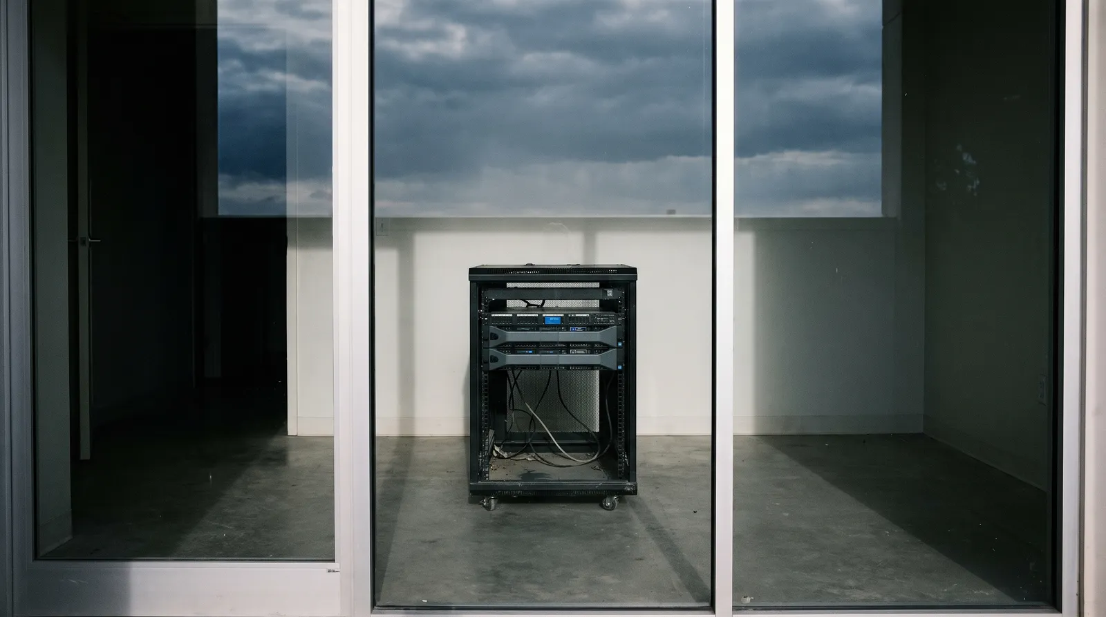 Small server rack alone in a glass-walled room, with cloudy sky reflected in the glass.